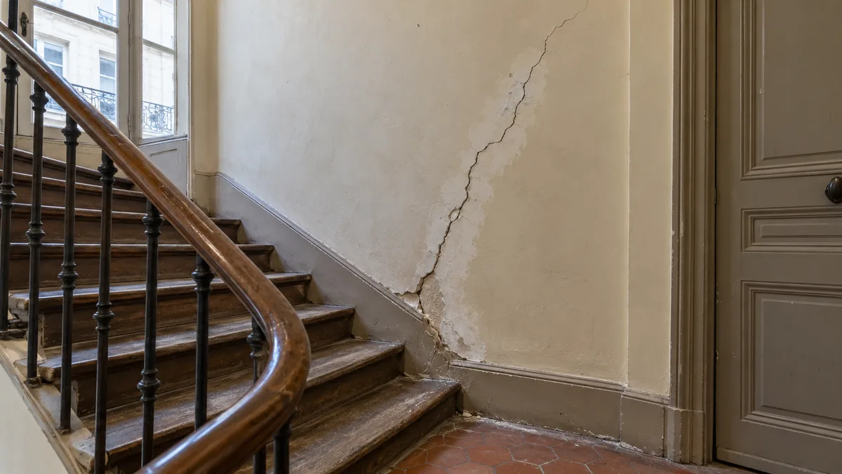 Cage d'escalier d'un immeuble ancien parisien, rampe en bois et tomettes au sol, où une longue fissure en diagonale part de la plinthe et monte dans l'enduit écaillé du mur