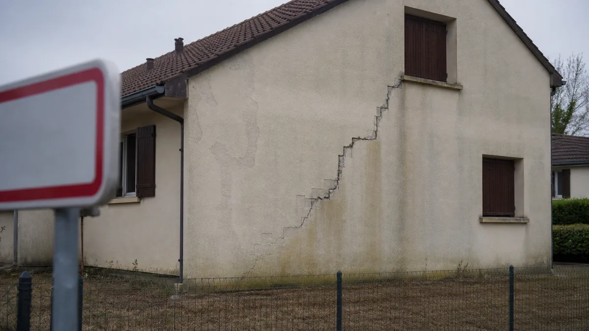 Pignon d'un pavillon de lotissement dont l'enduit est traversé par une longue fissure en escalier, ouverte et épaufrée, descendant de la fenêtre de l'étage jusqu'au bas du mur, panneau d'entrée de commune flou au premier plan, aucune personne dans le cadre