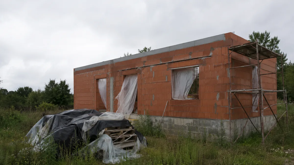 Chantier de maison individuelle à l'arrêt depuis des mois, échafaudage partiel et herbes hautes au pied des murs, aucune personne dans le cadre