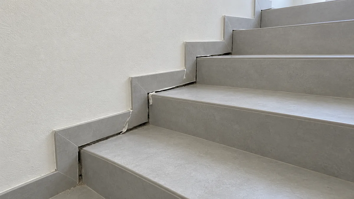 Photo d'un escalier droit en béton carrelé dans une maison : le long du mur blanc, la plinthe rampante est restée collée au mur alors que les marches sont descendues, laissant un jour sombre qui s'élargit de haut en bas jusqu'à la première marche