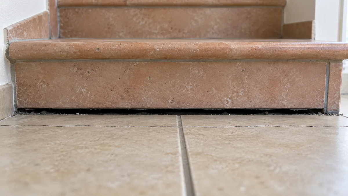 Photo au ras du sol du pied d'un escalier en béton carrelé : un jour sombre de près d'un centimètre s'est ouvert entre le dessous de la première marche et le carrelage du séjour, et deux carreaux voisins sont fendus sur une ligne parallèle à la marche