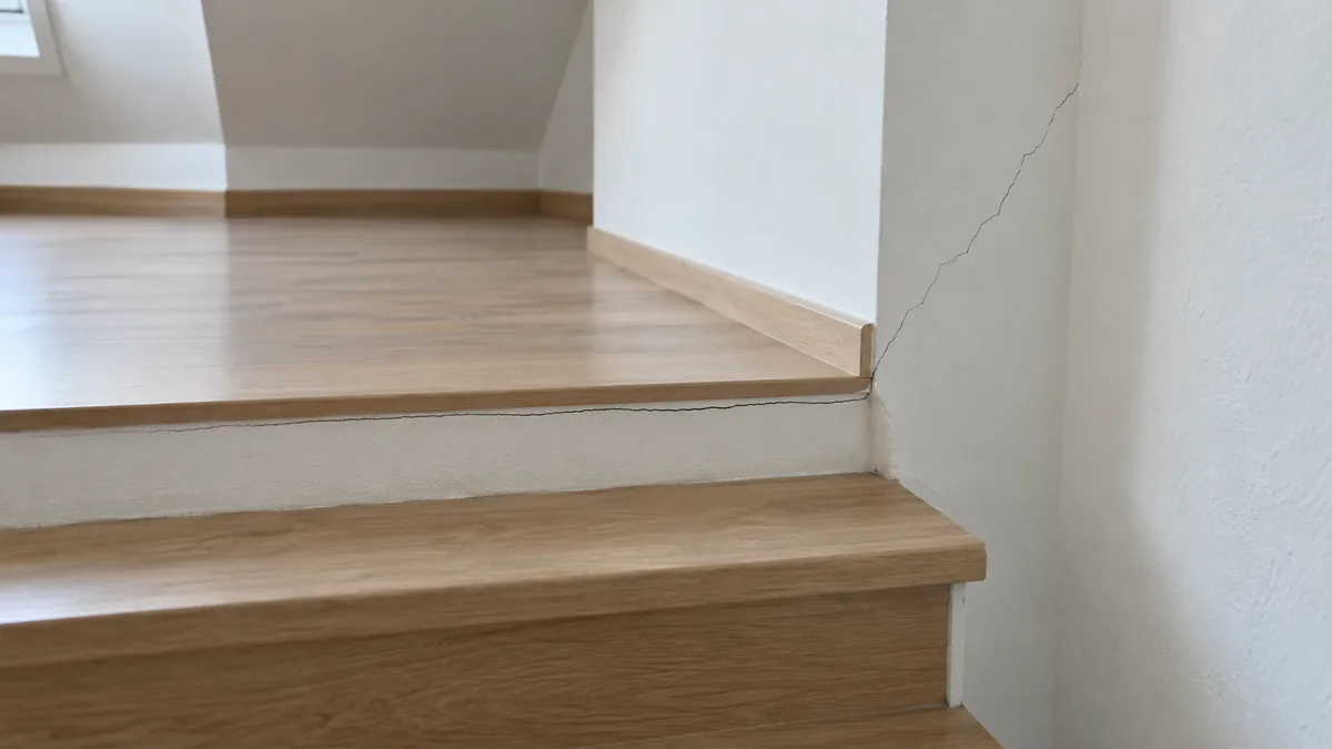 Photo du haut d'un escalier en béton vu depuis les marches : à l'arrivée sur le palier de l'étage, une fissure fine court le long du bord de la trémie entre la dernière marche et le plancher, puis remonte en biais sur le mur blanc depuis l'angle de l'ouverture