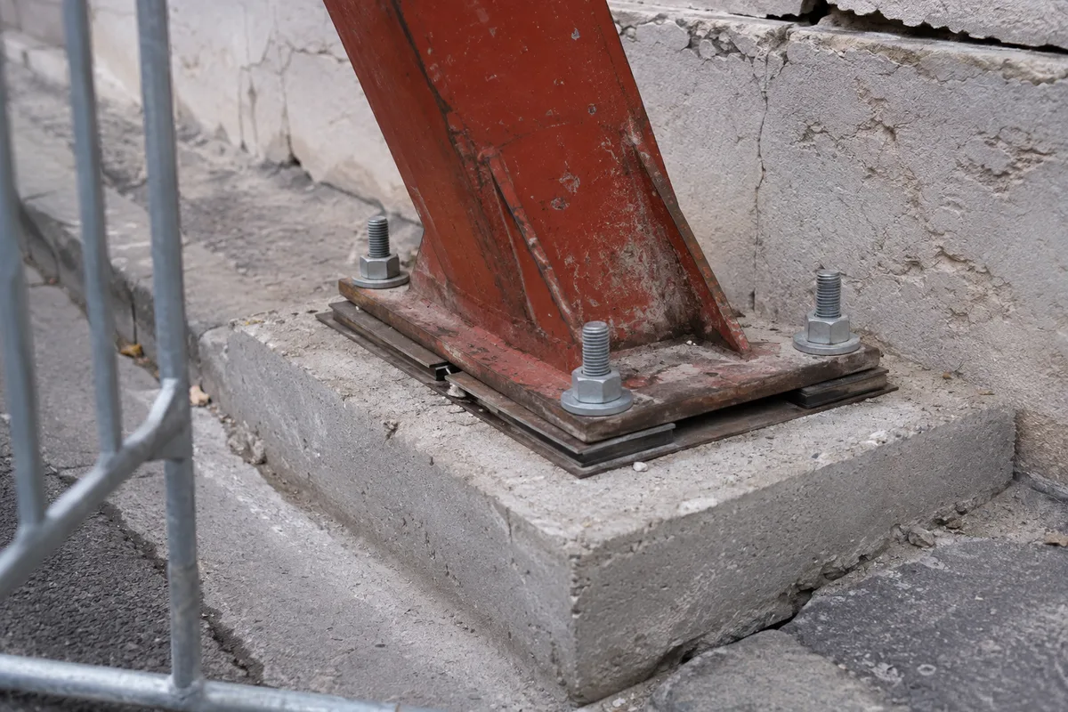Pied d'un buton de façade boulonné sur un massif de béton posé sur la chaussée, barrière de chantier au premier plan, aucune personne dans le cadre
