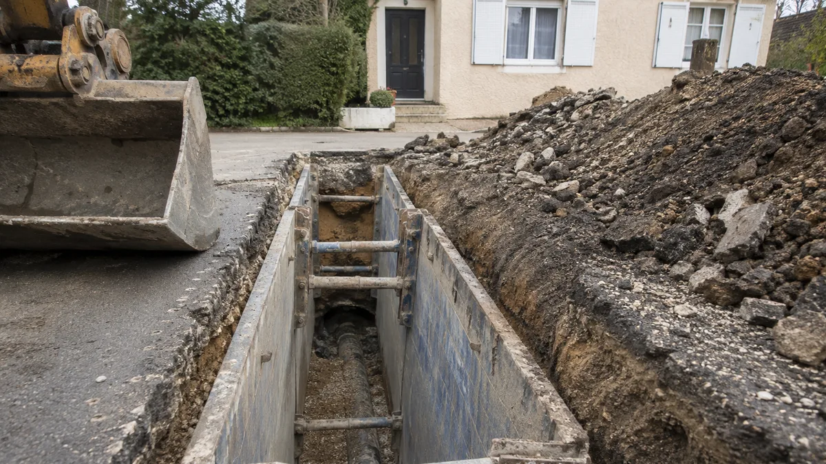 Tranchée de chantier ouverte dans une rue résidentielle bordée de maisons, blindages et engins en place, aucune personne dans le cadre
