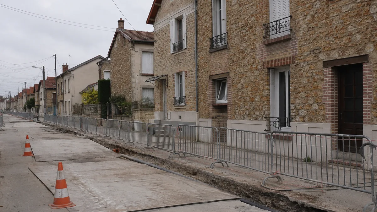 Tranchée de chantier de voirie ouverte le long d'une rue résidentielle, devant des pavillons mitoyens, sans personne dans le cadre