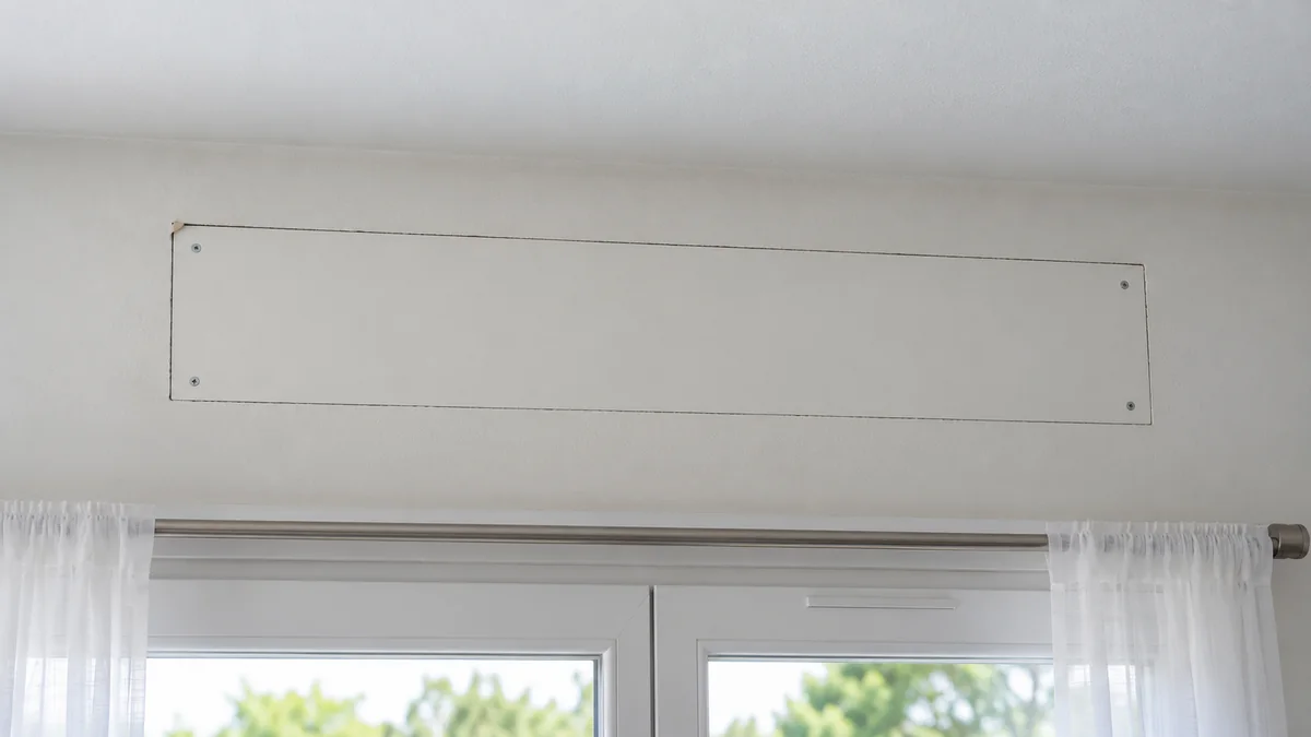 Intérieur d'une chambre, au-dessus d'une fenêtre : la trappe de visite rectangulaire du coffre de volet roulant est entourée d'une fissure fine dans le plâtre qui suit exactement son contour, avec un petit éclat de peinture à un angle