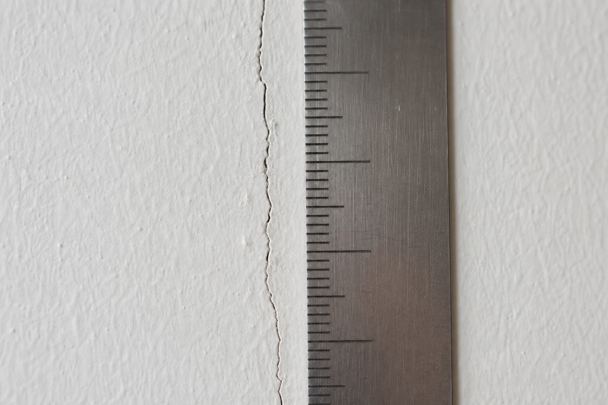 Gros plan sur une fissure fine de mur intérieur avec un réglet gradué posé à côté pour donner l'échelle de la photo
