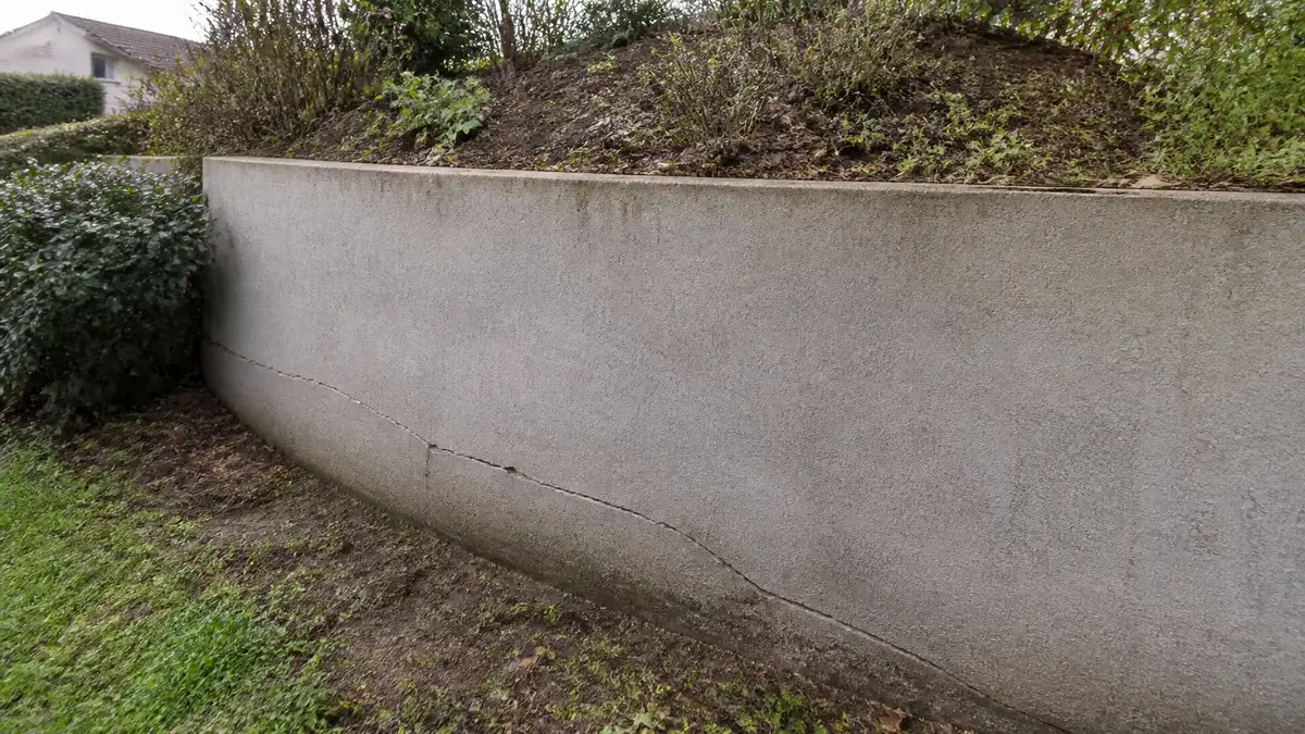 Mur de soutènement enduit en fond de jardin résidentiel avec une fissure diagonale nette et un léger bombement visible en partie basse, végétation en haut du talus