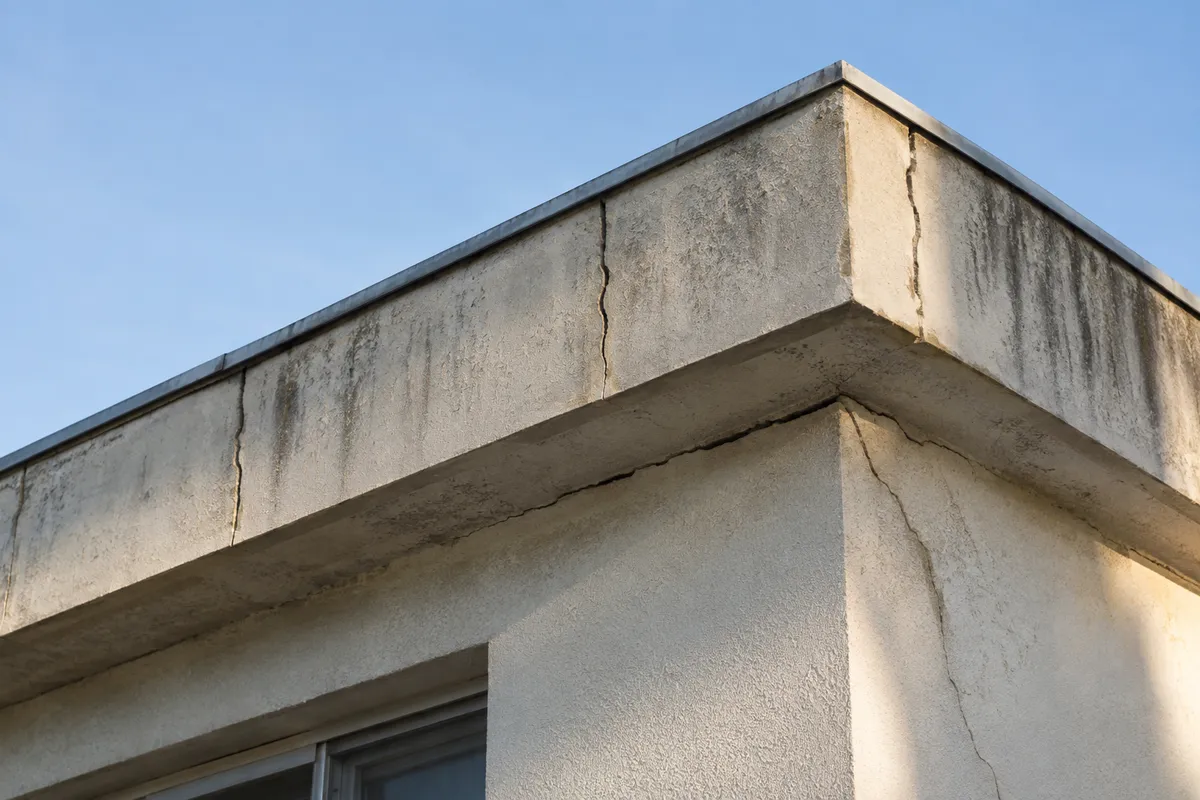 Angle d'un pavillon à toit plat des années 1970 en Île-de-France, acrotère en béton coupé de fissures verticales régulières et fissure horizontale sous la dalle de toiture qui descend en biais à l'angle de la façade, enduit taché par les coulures, aucune personne