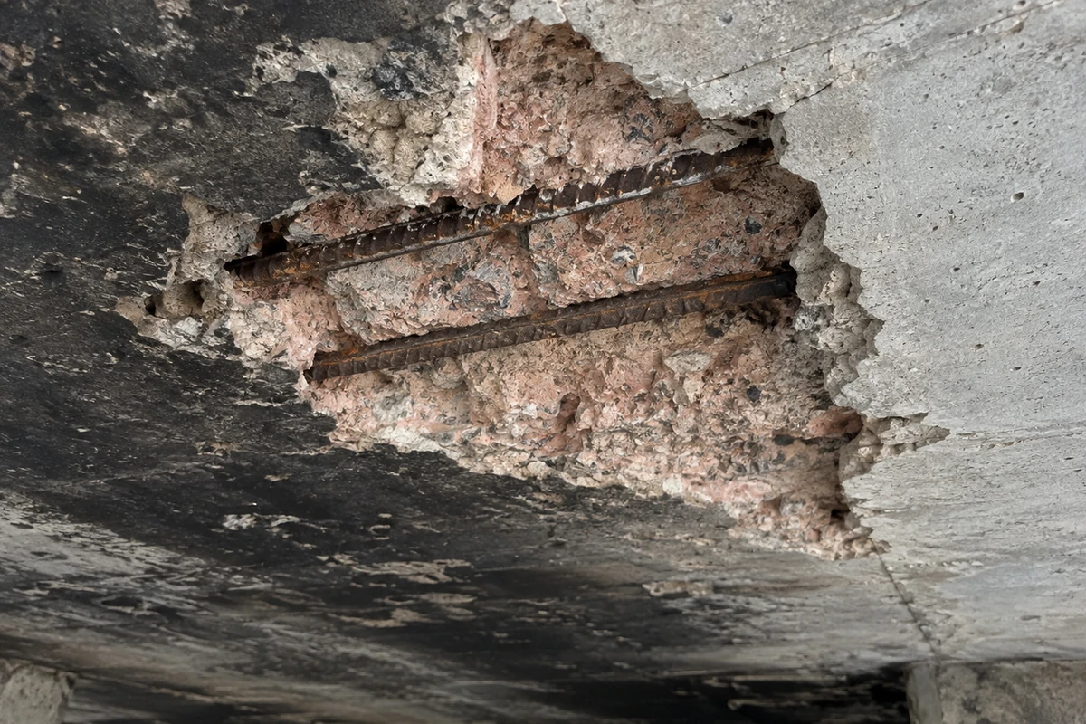Sous-face d'une dalle béton après incendie, parement éclaté sur plusieurs dizaines de centimètres laissant apparaître les armatures nues et du béton teinté rose, aucune personne dans le cadre