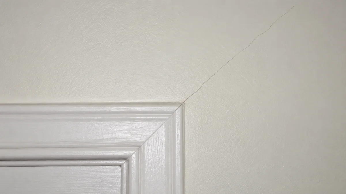Vue rapprochée d'un mur intérieur peint en blanc cassé avec une fissure fine qui part de l'angle supérieur d'un encadrement de porte