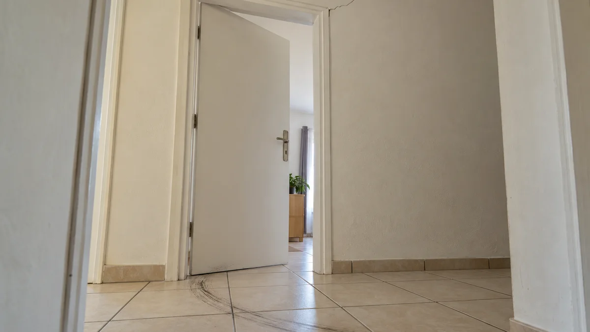 Porte intérieure entrouverte dont le battant frotte sur le carrelage, laissant un arc de rayures au sol, sous une fissure fine qui part de l'angle de l'huisserie