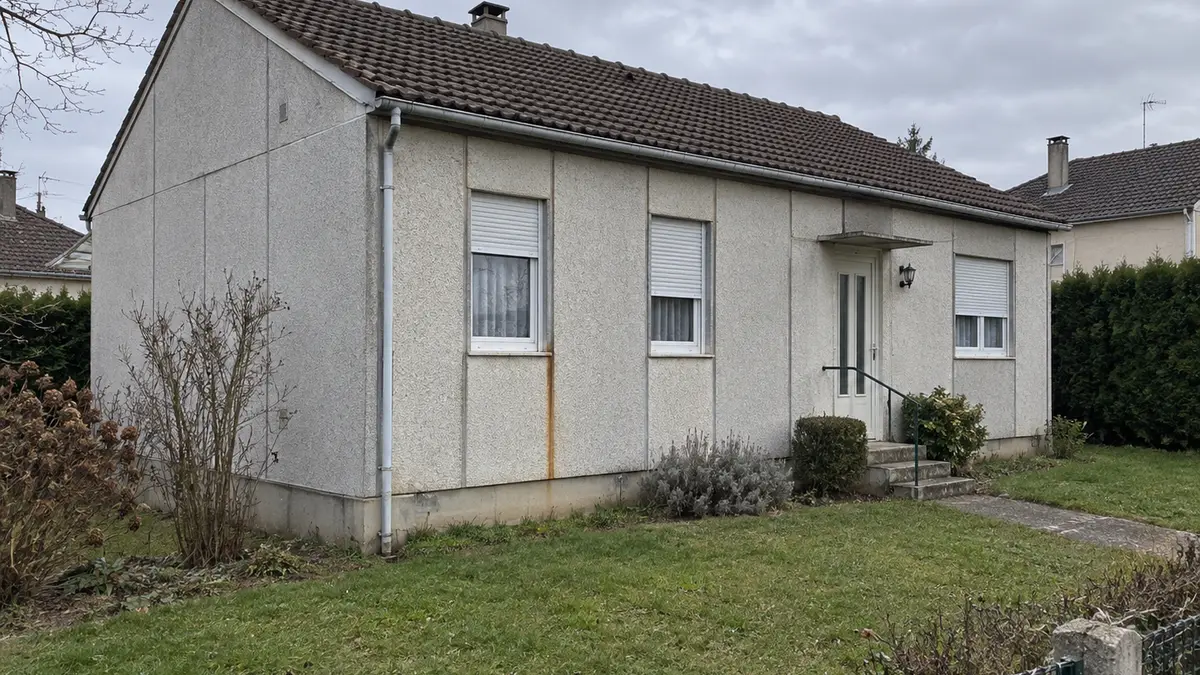 Maison Phénix de plain-pied des années 1970 en banlieue parisienne, façade en panneaux de béton préfabriqués aux joints verticaux réguliers, trace de rouille sous un joint, jardin et clôture basse, aucune personne