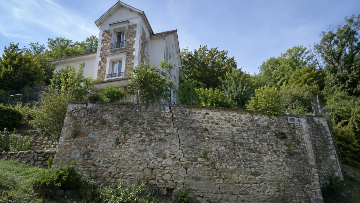Maison implantée sur un terrain en pente avec un mur de soutènement en moellons bombé et fissuré, aucune personne dans le cadre