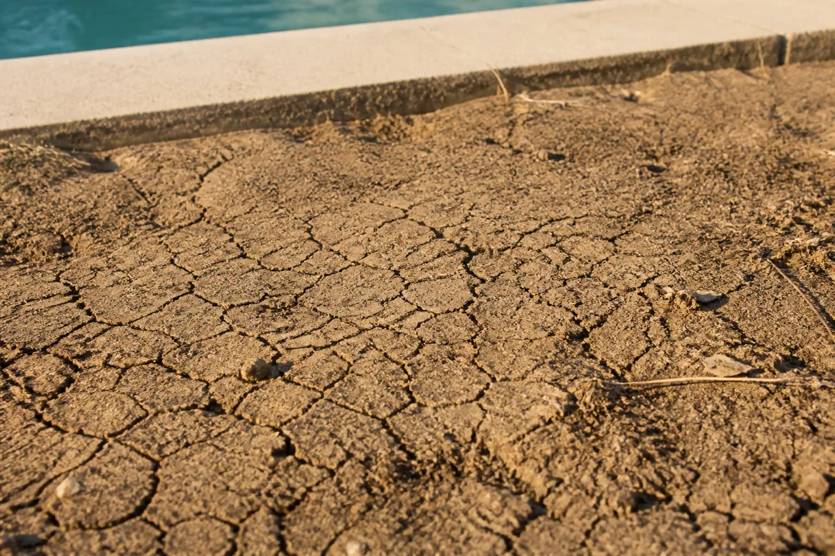 Sol argileux craquelé en surface par la sécheresse estivale, à quelques dizaines de centimètres de la margelle d'une piscine, aucune personne dans le cadre