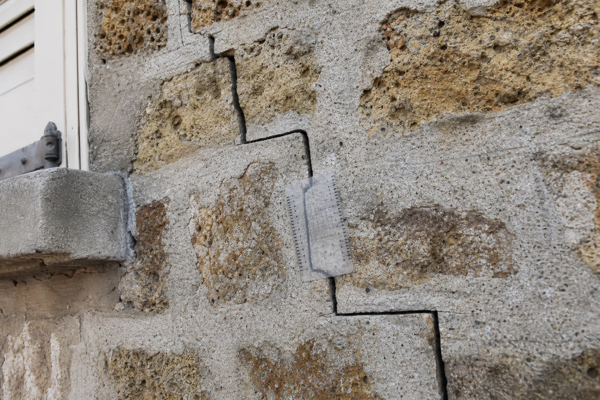 Fissure diagonale en escalier sur un mur de façade francilien, avec une jauge de mesure posée à côté pour l'échelle