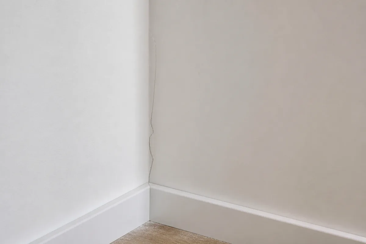 Fissure fine à l'angle d'un mur fraîchement enduit dans un logement neuf, traces de poussière de chantier encore visibles au sol