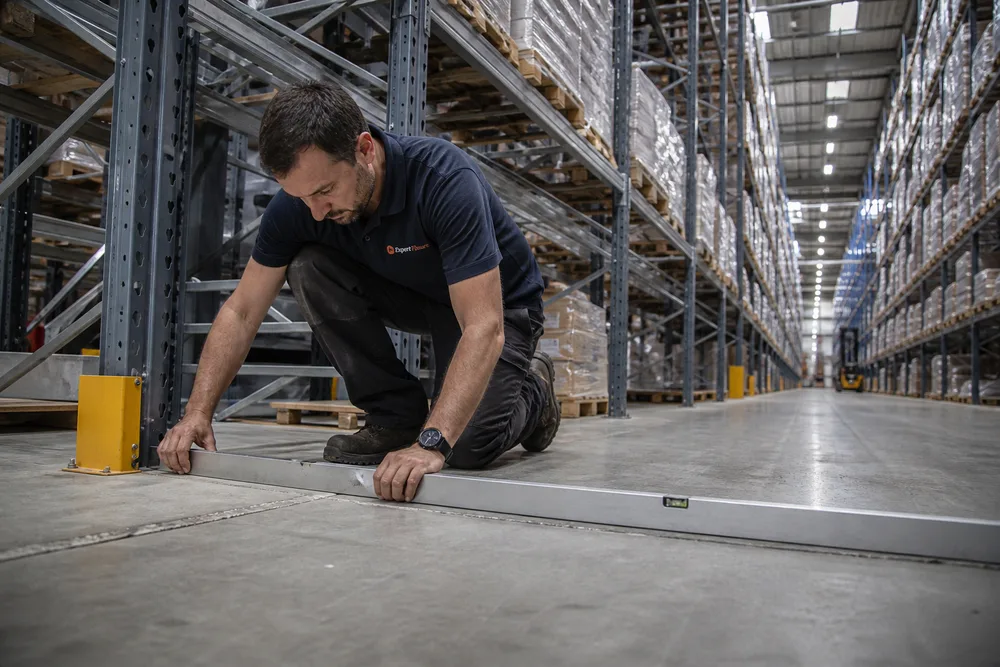 Ingénieur Expert Fissure agenouillé au pied d'un rack de stockage, contrôlant la planéité du dallage à la règle métallique dans un entrepôt logistique