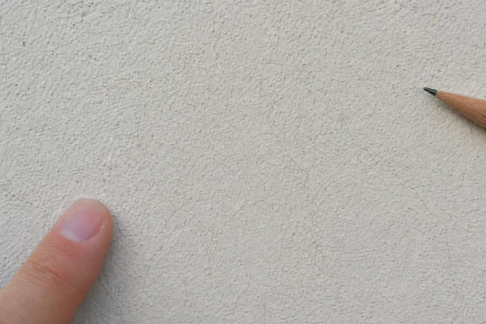 Réseau de microfissures fines en toile d'araignée sur un enduit de façade, doigt et crayon pour l'échelle